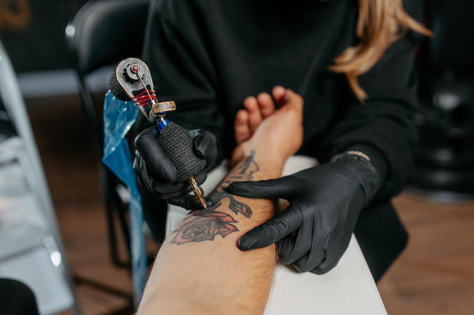 Tattoo artist wearing black gloves tattoos a forearm with a blue machine