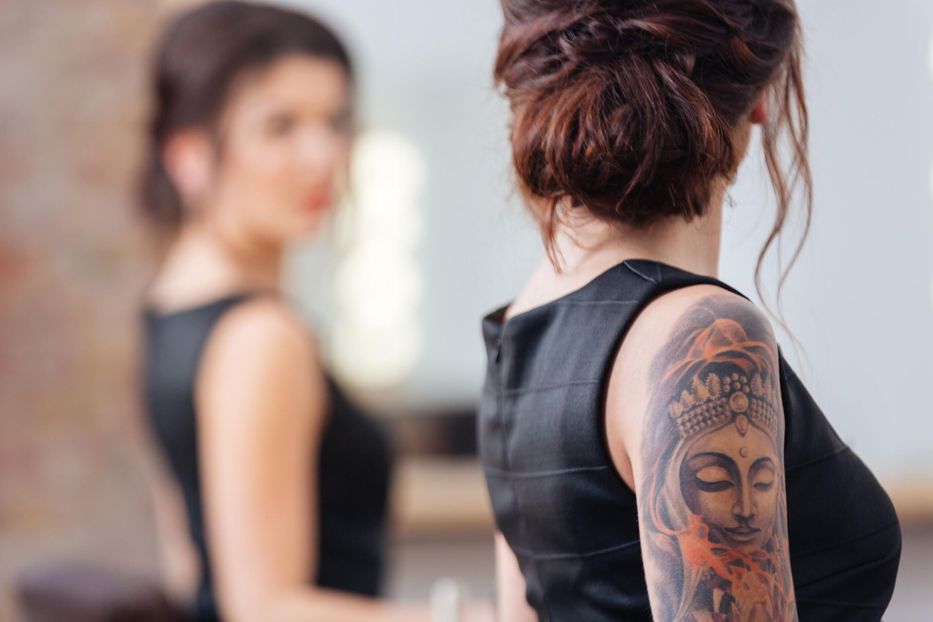 Woman with tattooed arm in black sleeveless top, standing in a studio with a blurred reflection in the background