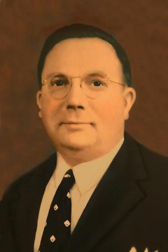Formal portrait of a man in a dark suit and tie against a warm brown background