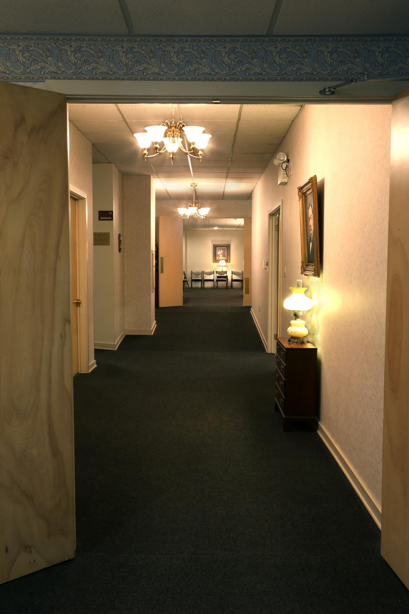 Dim carpeted hallway with white walls, ceiling lights, framed art, and a lit lamp on a small table.