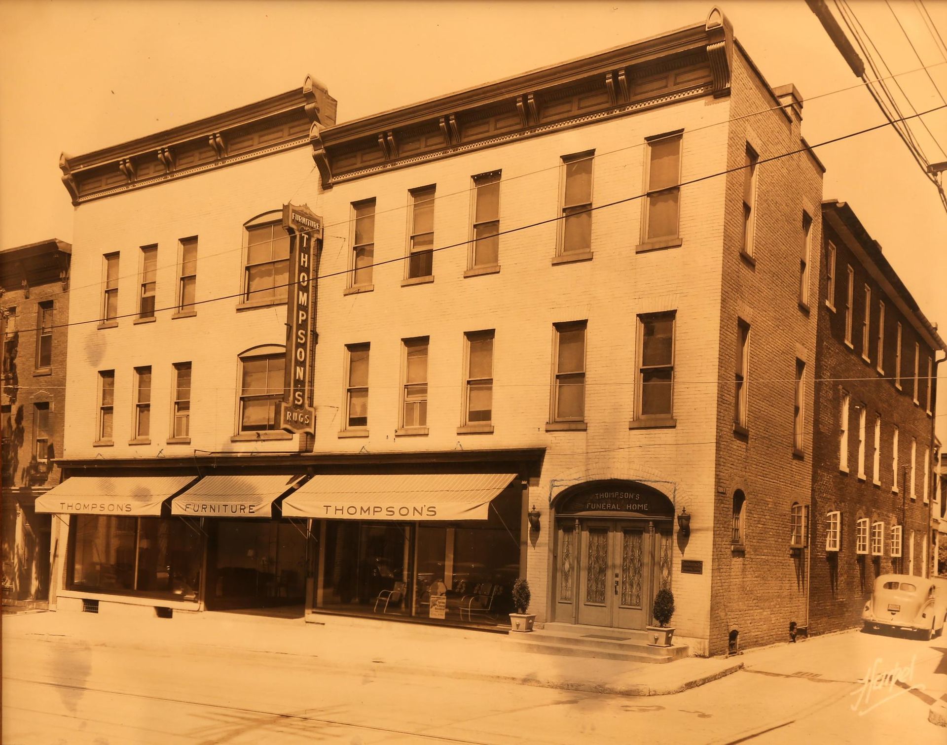 Sepia-toned street view of a corner brick building with storefront awnings and arched entrances.
