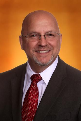 Portrait of a smiling bald man in a dark suit, white shirt, and red tie against an orange background