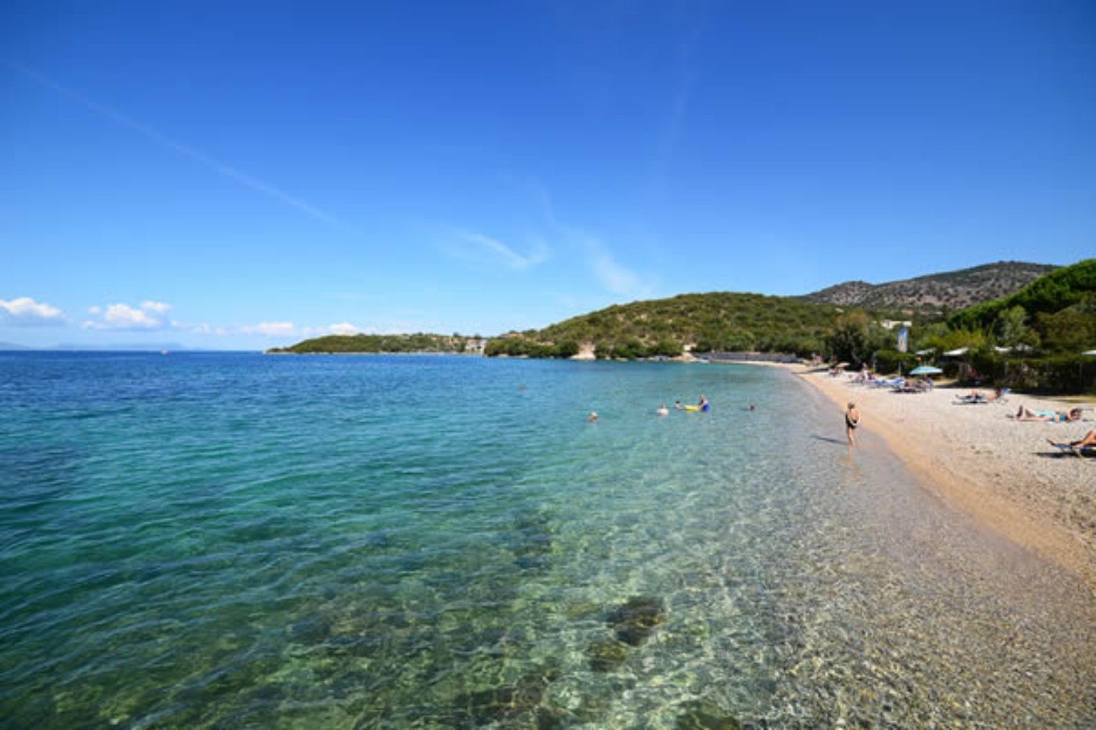 Clear turquoise beach with a sandy shore, calm water, and green hills under a blue sky