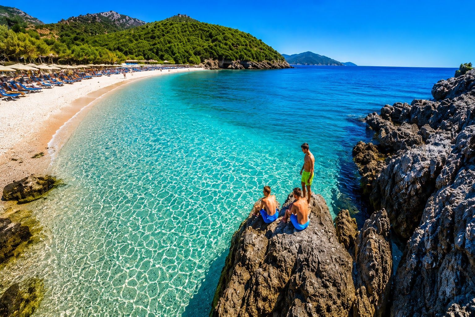 People sitting on sunlit rocks beside a turquoise tropical bay and sandy beach