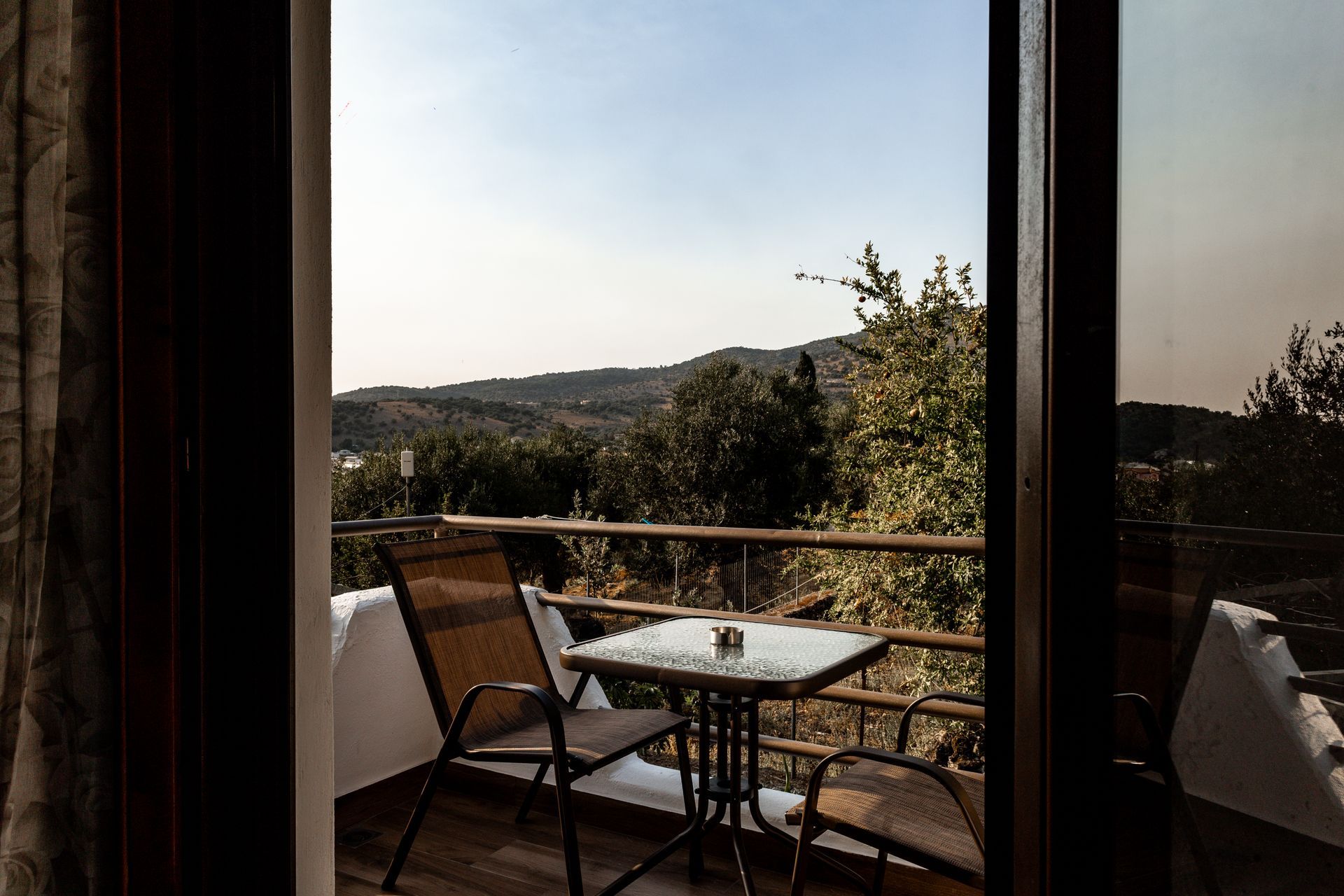 Balcony table and chairs overlooking a sunlit hillside landscape through glass doors