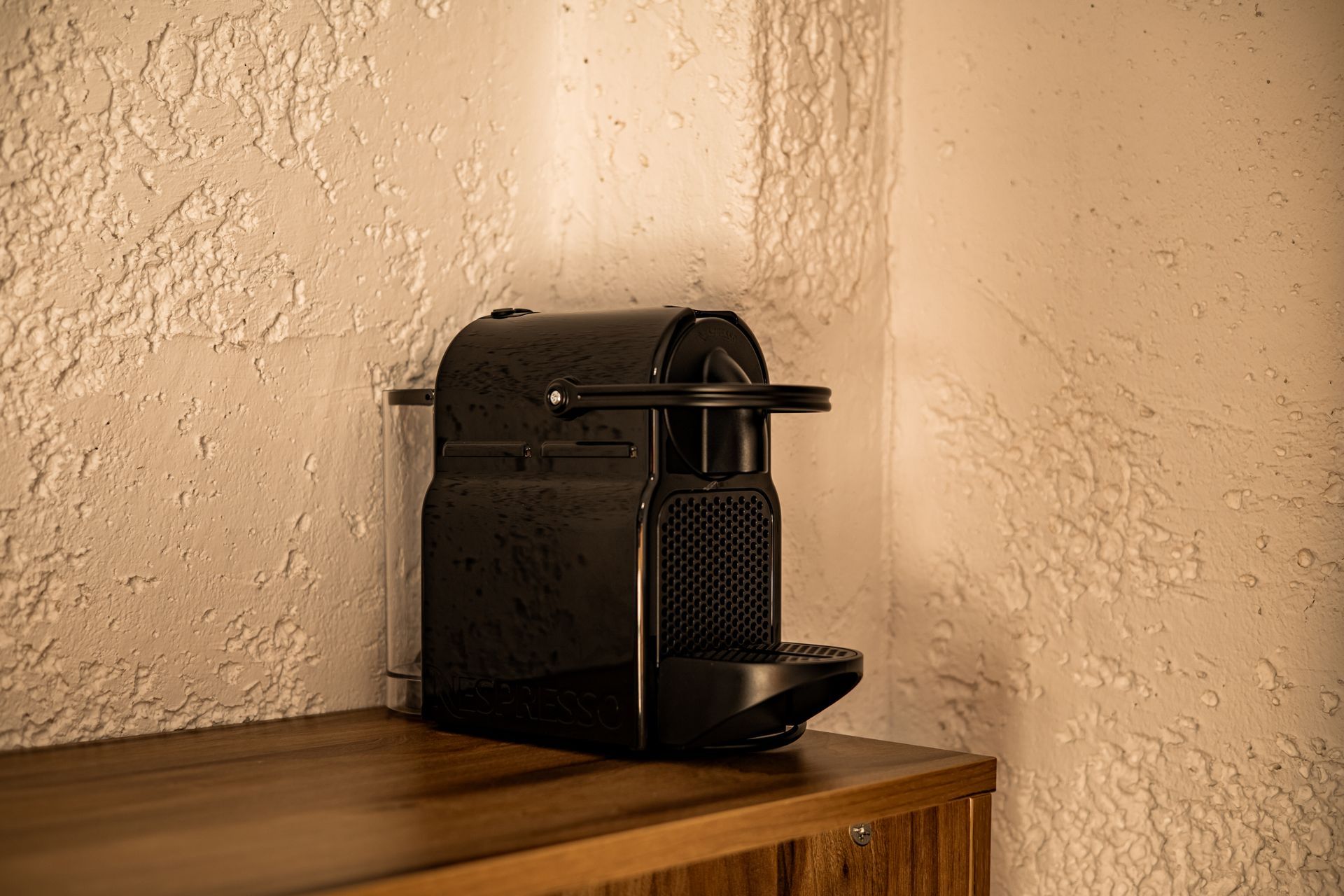 Black Nespresso-style coffee machine on a wooden shelf against a textured beige wall