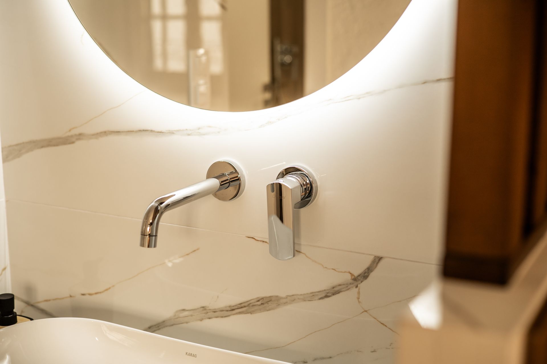 Modern bathroom sink with round mirror, chrome wall faucet, and beige marble tiles