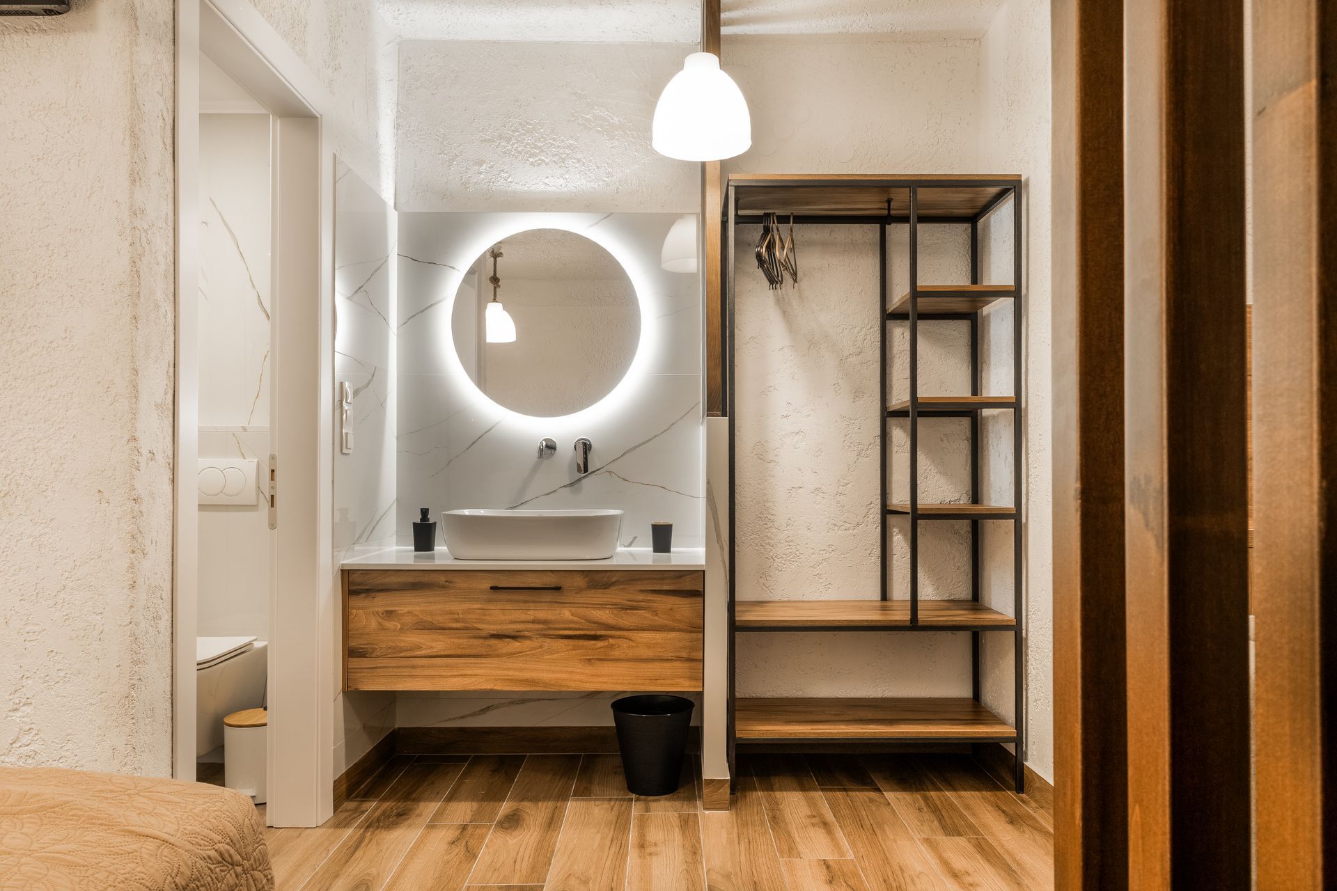 Modern bathroom with wood vanity, round lit mirror, white walls, and open shelving.