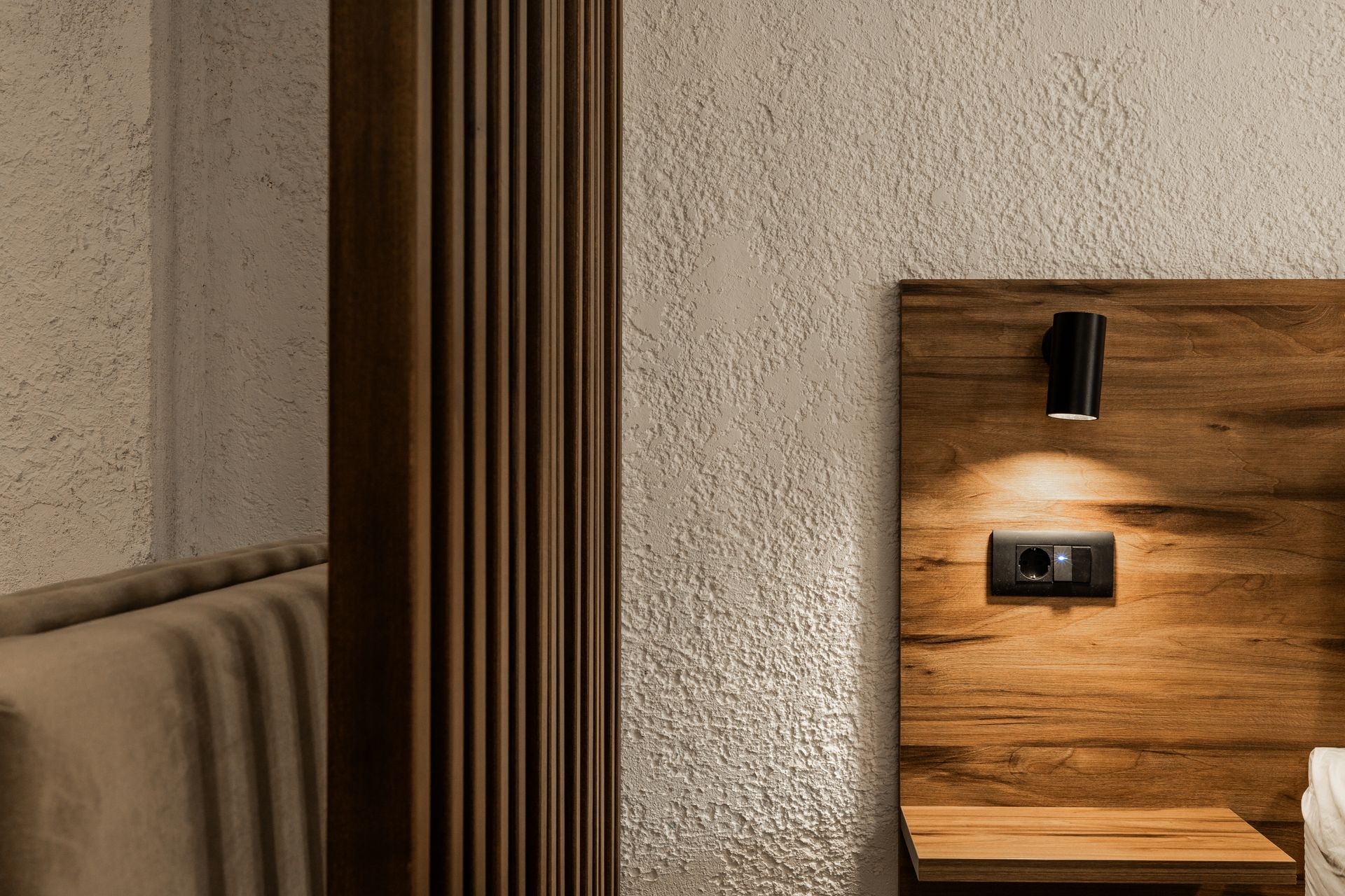 Modern bedroom corner with textured walls, wood accent panel, bedside light, and a bed edge