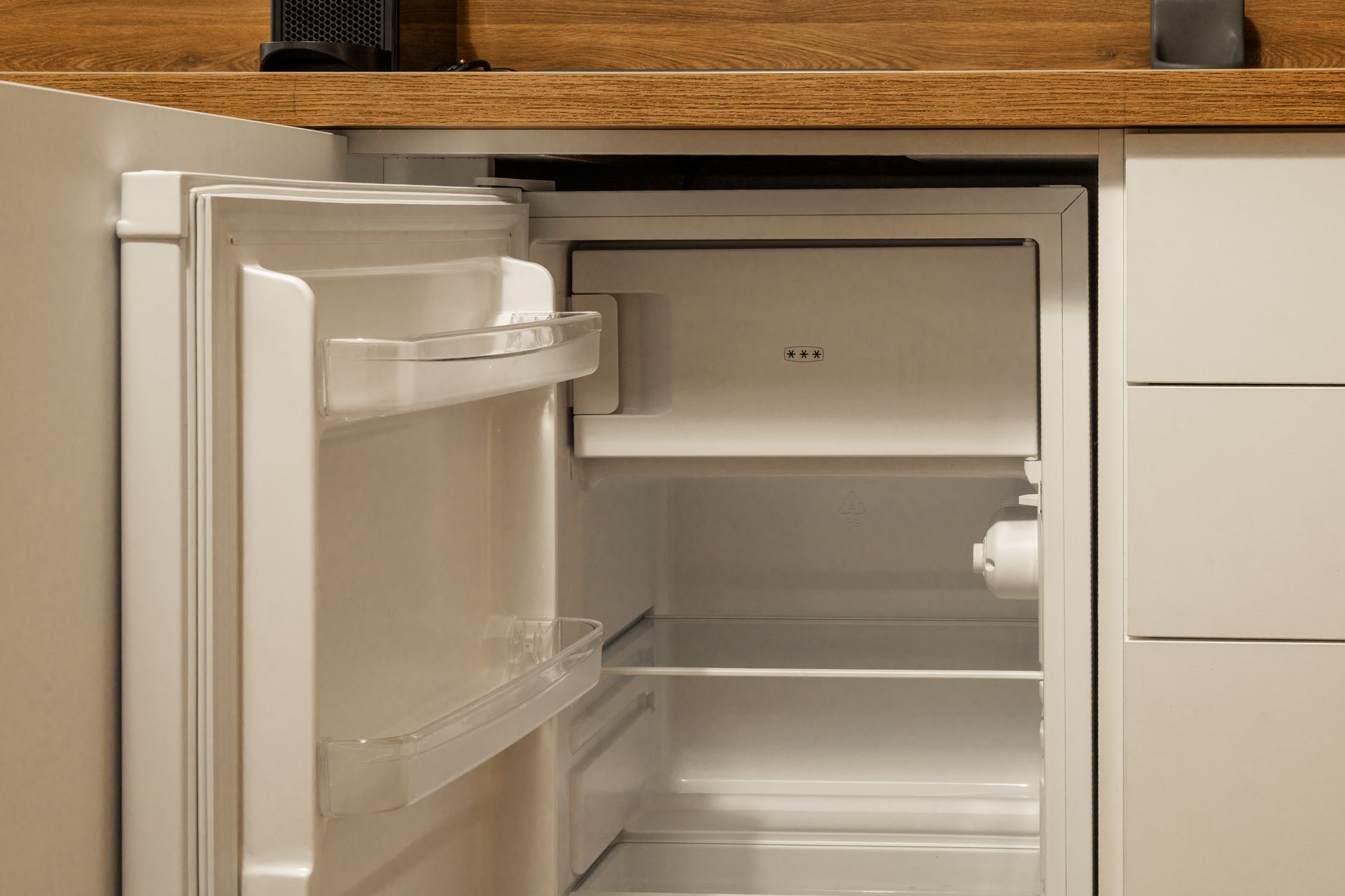 Open mini fridge under wooden counter, door ajar, with white shelves and an empty interior.