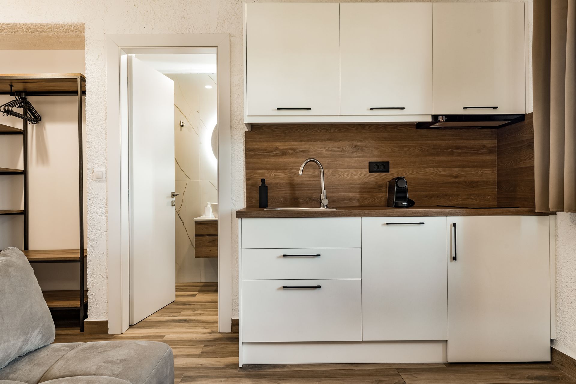 Modern kitchenette with white cabinets, wood backsplash, sink, and open doorway in a small apartment.
