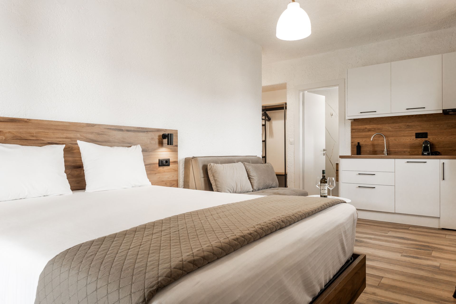 Modern bedroom with wooden bed, white bedding, and compact kitchen area in the background