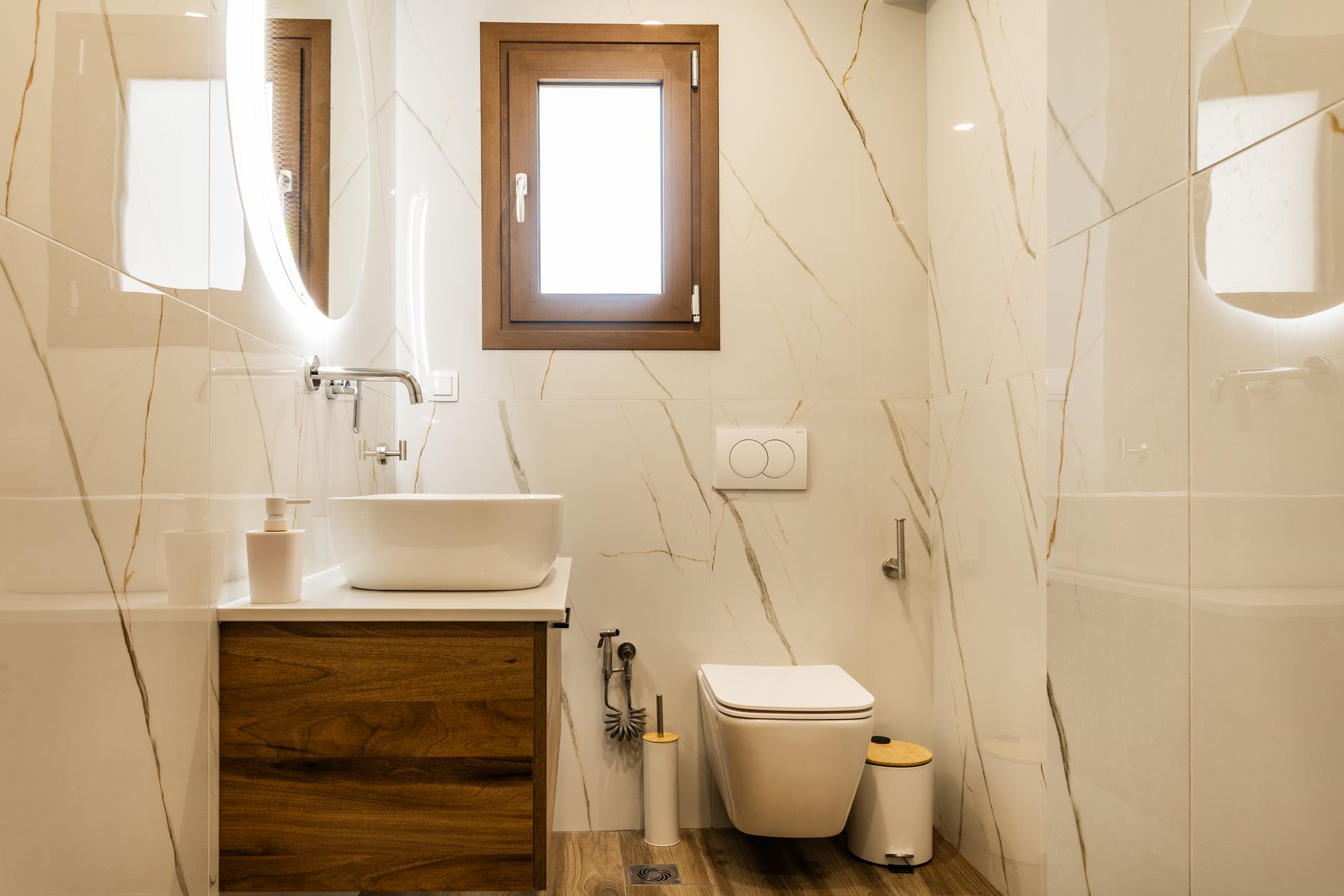 Small modern bathroom with sink, toilet, mirror, and wall sconce lighting on beige marble-patterned tiles