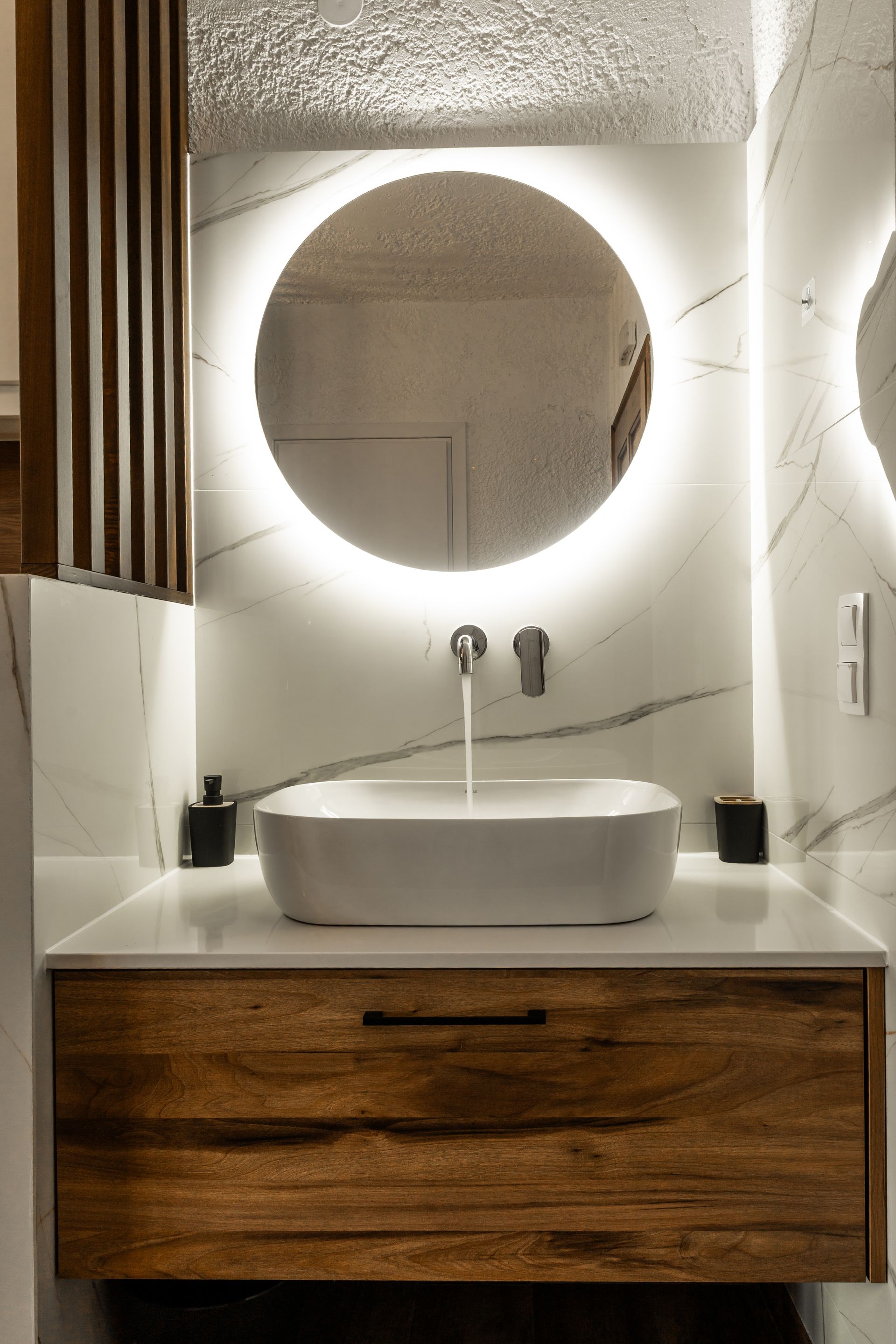 Modern bathroom vanity with round backlit mirror, white sink, and wooden cabinet drawers.