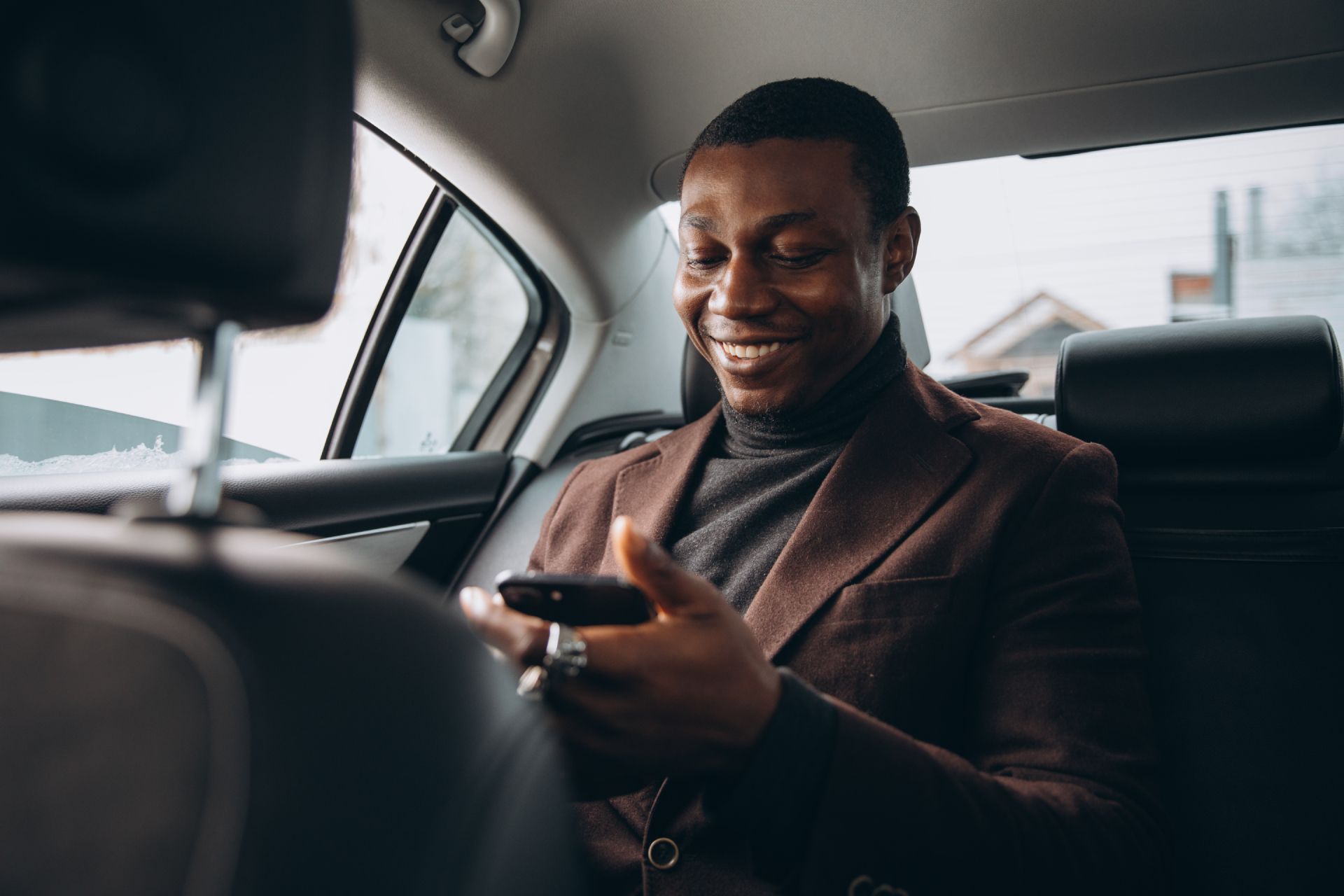 Smiling man in a brown blazer using a smartphone in the back seat of a car