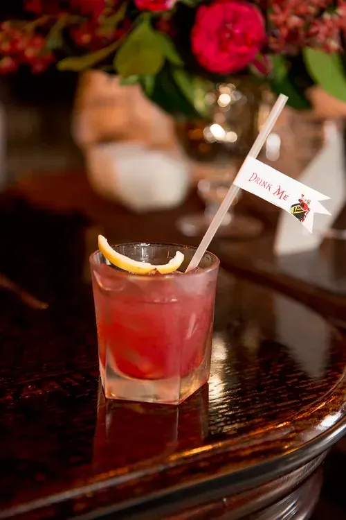 Pink cocktail with lemon garnish and a straw on a wooden table, with flowers blurred in the background