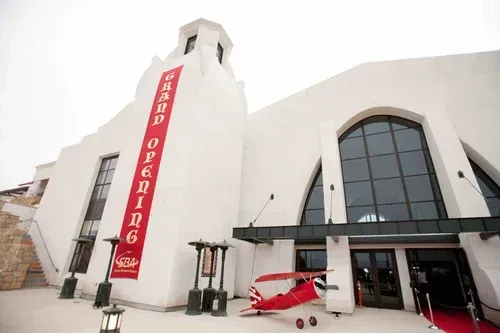 Modern white building with a tall red vertical sign and arched glass entrance, a small red airplane outside