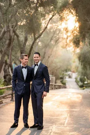 Two men in navy suits stand on a sunlit tree-lined path, smiling at each other.