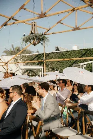 Guests seated outdoors under white umbrellas at a formal event with a wooden canopy overhead