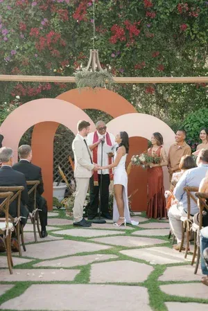 Outdoor wedding ceremony under floral arch with couple exchanging vows, guests seated on both sides.