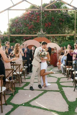 Bride and groom kissing in a garden aisle during an outdoor wedding ceremony, guests applauding on both sides