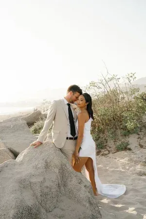 Couple embracing on a beach beside a large rock, wearing light-colored formal attire at sunset.
