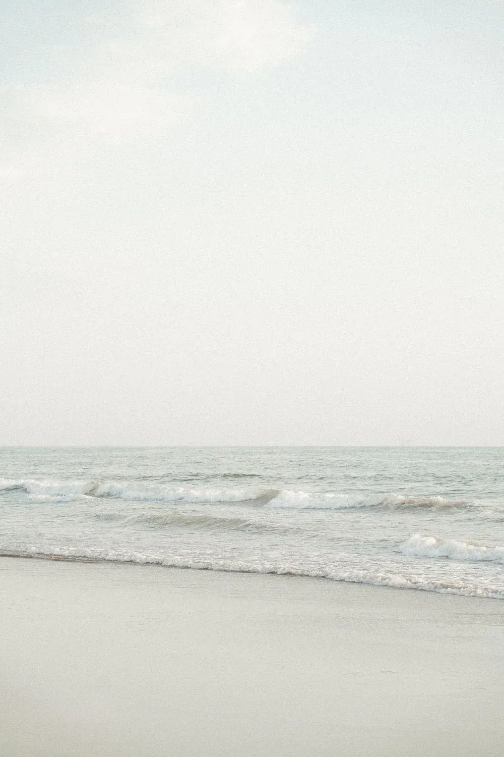 Calm beach with pale sand, gentle waves, and a bright overcast sky
