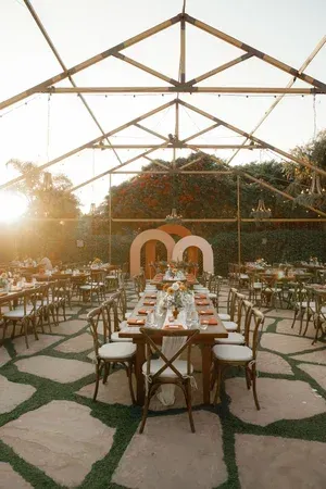 Outdoor wedding reception with long tables, white chairs, and a wooden frame at sunset