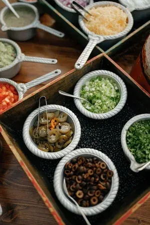 Assorted chopped toppings in small bowls with spoons on a dark tray, viewed from above