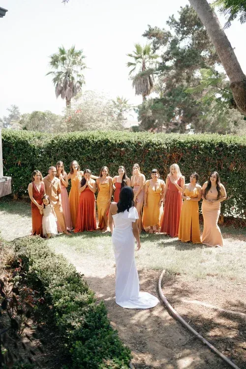 Wedding party in orange and white dresses stands outdoors facing a person in white.