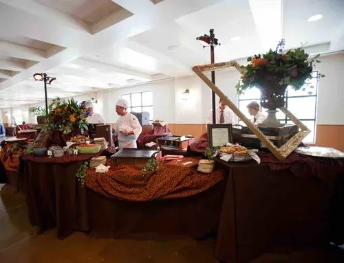Buffet table with floral arrangements and desserts in a bright banquet room, with a person standing behind it