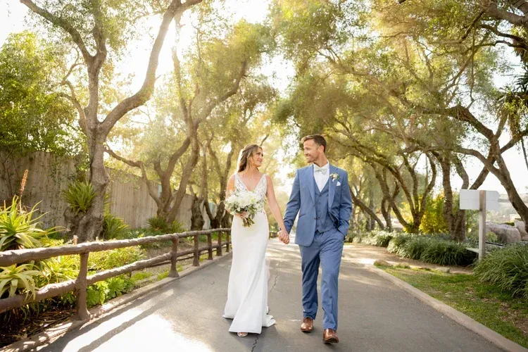 Couple in wedding attire walking hand in hand along a sunlit garden path