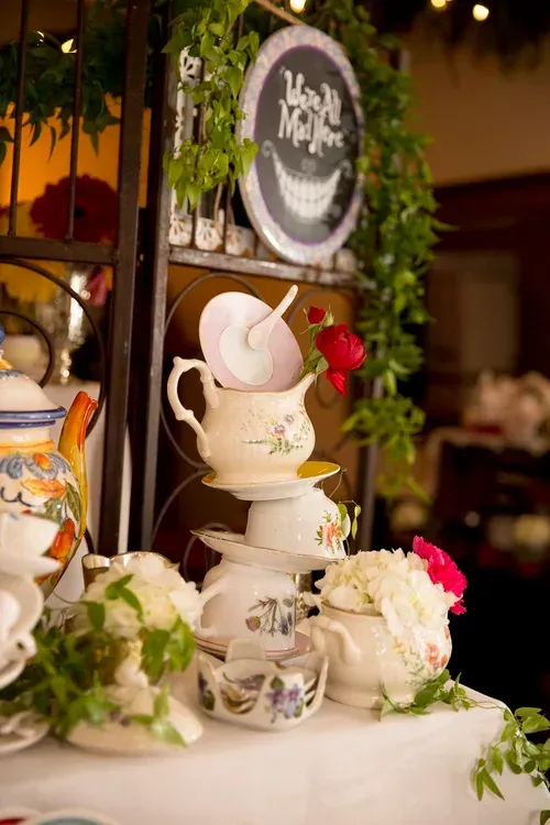 Elegant dessert table with stacked teacups, white cakes, flowers, and a chalkboard sign with greenery