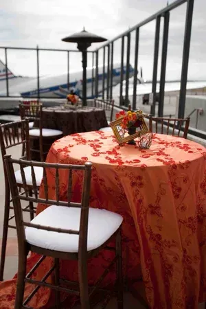 Outdoor café terrace with red floral tablecloth, wooden chairs, and a harbor view under cloudy skies