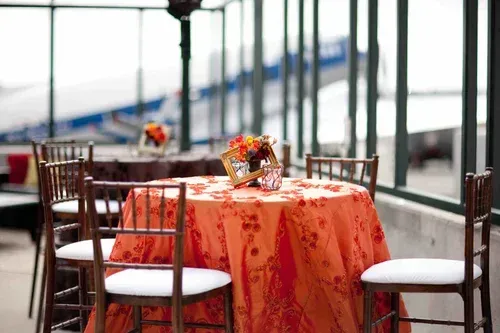 Outdoor café table with orange tablecloth, floral centerpiece, and chairs by a railing, overlooking a blurred bridge