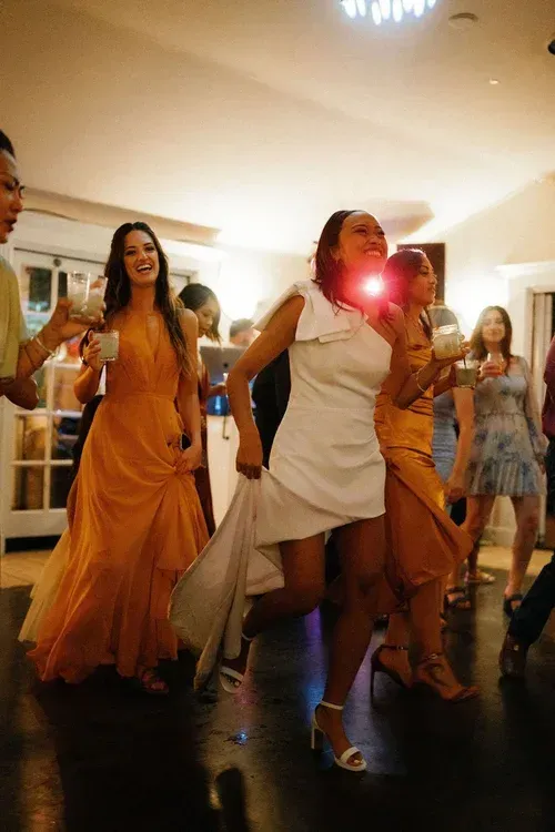 Three women in flowing dresses dance at a lively indoor party, with a bright flash at center.