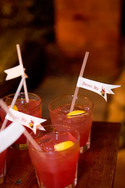 Three pink cocktails with lemon slices and white paper straws on a wooden table, one with a “Drink Me” tag
