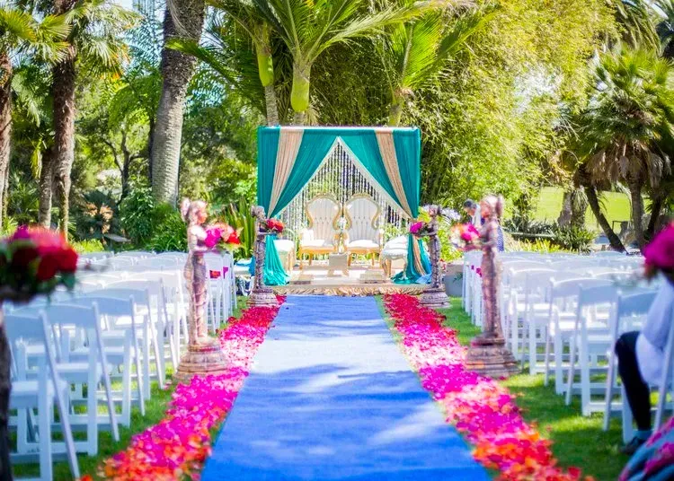 Outdoor wedding aisle with white chairs, blue runner, and teal draped altar under palm trees