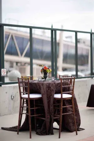 Small café table with dark tablecloth, flowers, and two wooden chairs by a glass window.