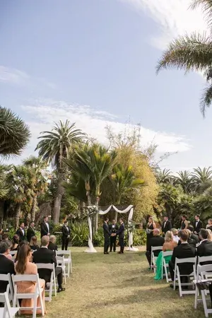 Outdoor wedding ceremony on a grassy lawn with guests seated among palm trees and white chairs