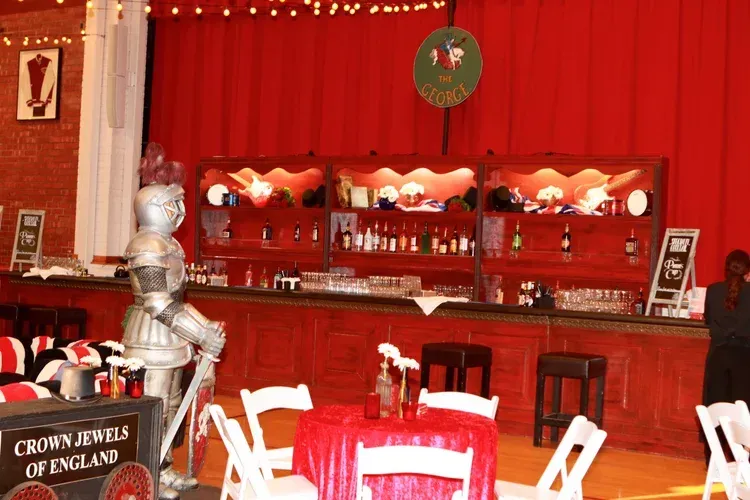 Red-lit bar with shelves, bottles, and white chairs beside a silver knight statue
