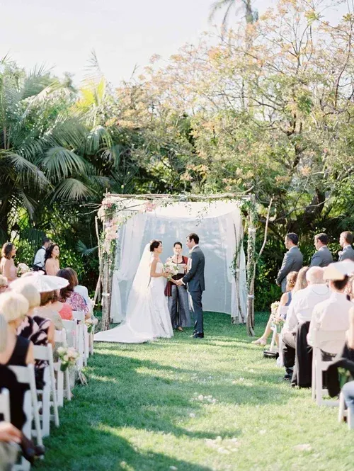Outdoor wedding ceremony under a floral arch, with bride and groom facing each other along a garden aisle