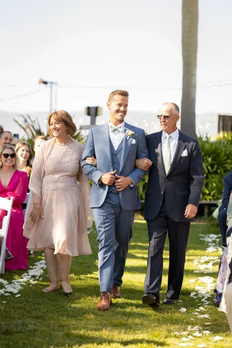 Three people in formal attire walking outdoors at a garden event, arm in arm.