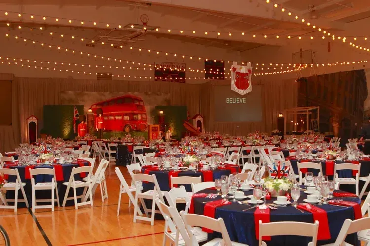 Decorated banquet hall with round tables, red-and-blue tablecloths, white chairs, and string lights overhead