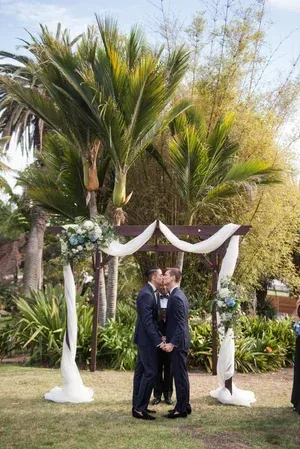 Two men in suits standing under a white draped wedding arch in a garden with palm trees
