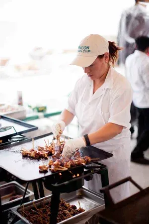 Person wearing a cap grills skewers at a food stall.