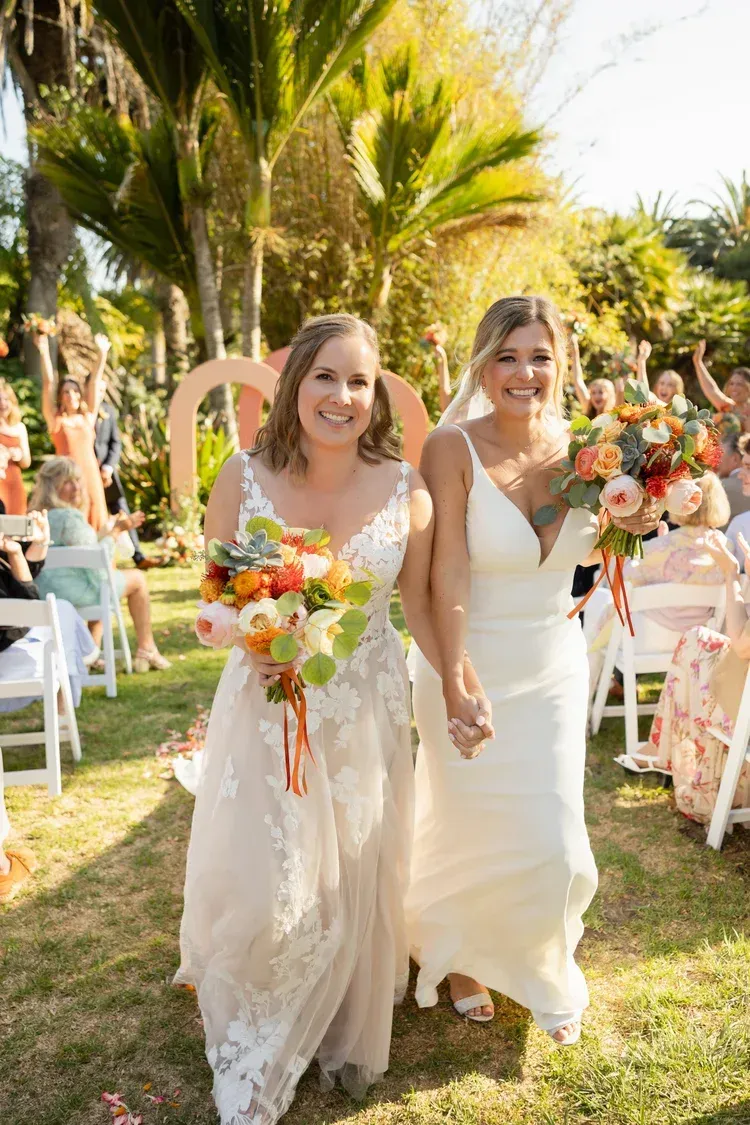 Two brides in white dresses walking hand in hand at an outdoor wedding with bouquets and palm trees