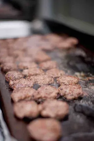 Several hamburger patties cooking on a smoky grill, with one spatula in the background