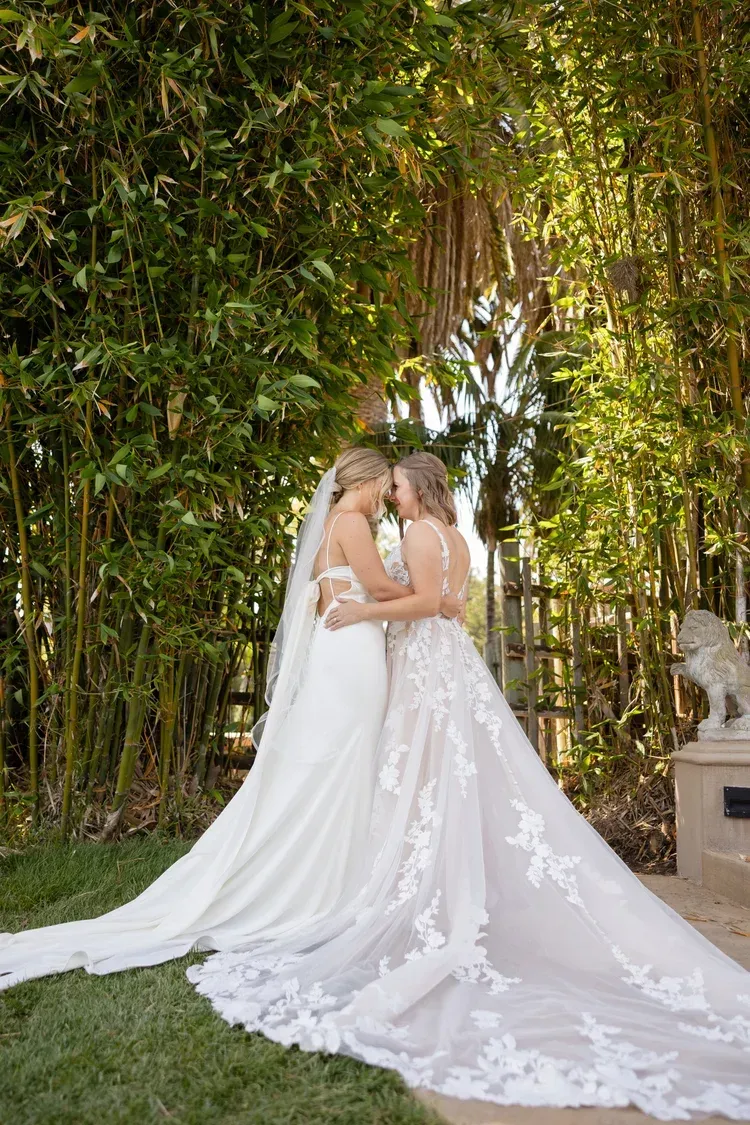 Two brides in white wedding gowns embracing under a leafy bamboo archway