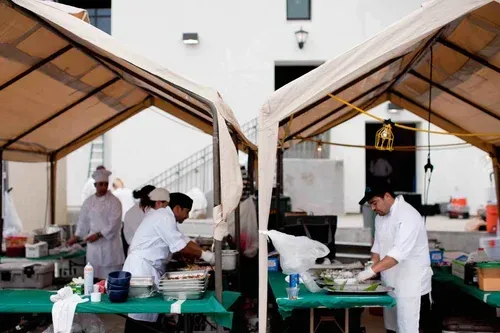 Chefs preparing food at outdoor catering stations under beige canopies.